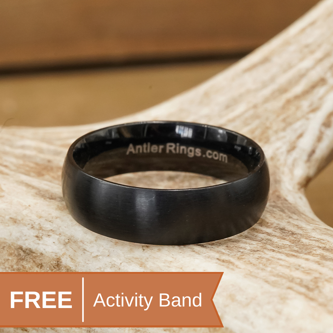 A black titanium antler ring called The Shield of Faith, with AntlerRings.com engraved inside and a hammered finish, rests on an antler. An orange banner in the corner reads FREE Activity Band.
