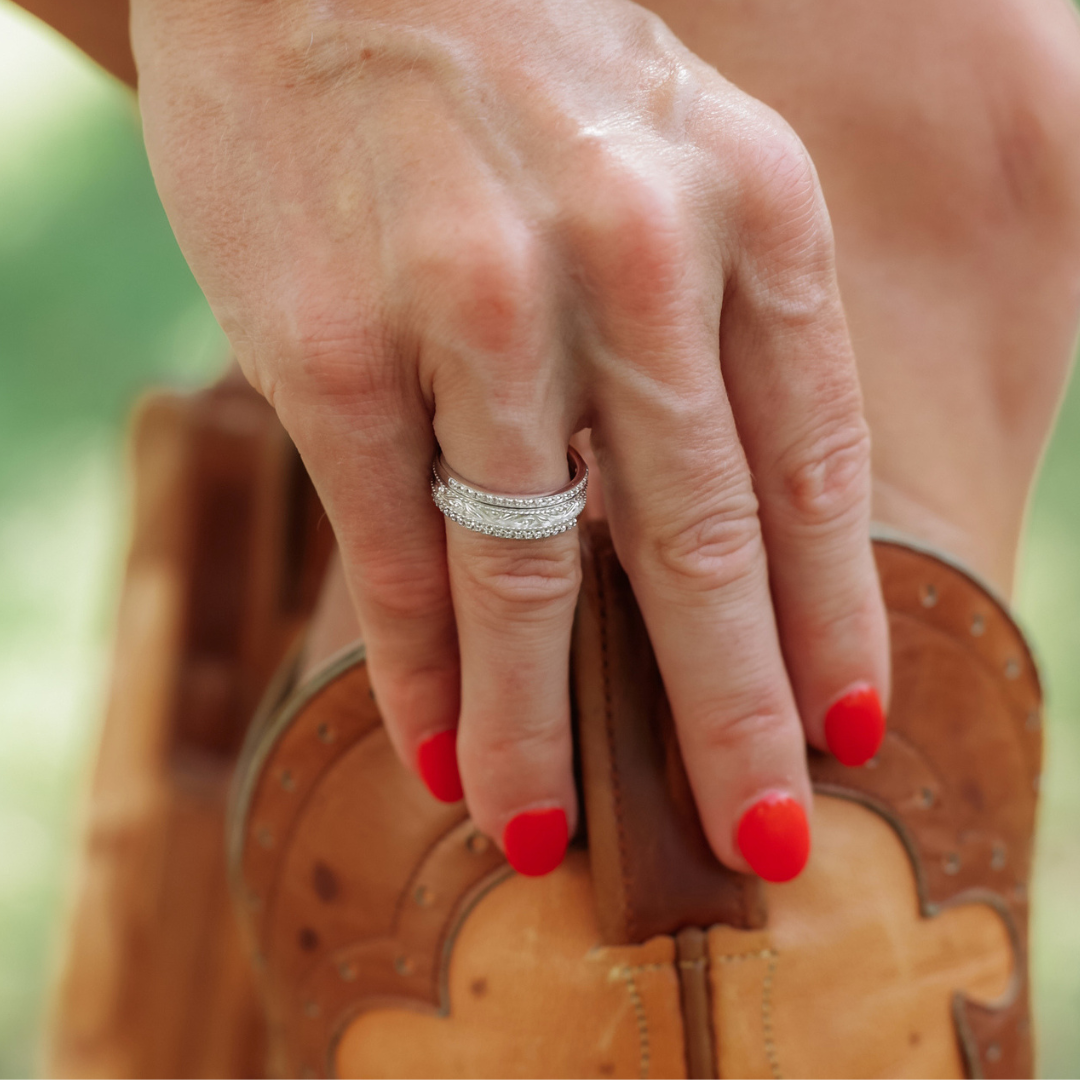 A hand with red painted nails rests on a leather saddle, wearing three stacked rings from The Addison Stacker Set on the ring finger. These elegant bands feature brilliant cubic zirconia that catches the light beautifully.