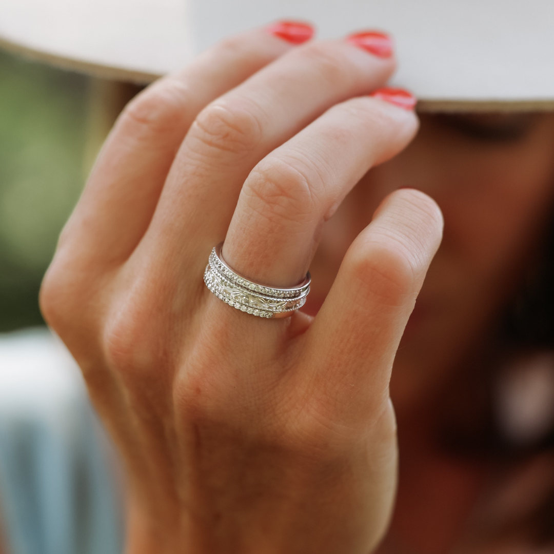 A person with coral-painted nails wears The Addison Stacker Set—multiple cubic zirconia stacking bands—while holding a white hat that partially covers their face. The background is blurred and set outdoors.