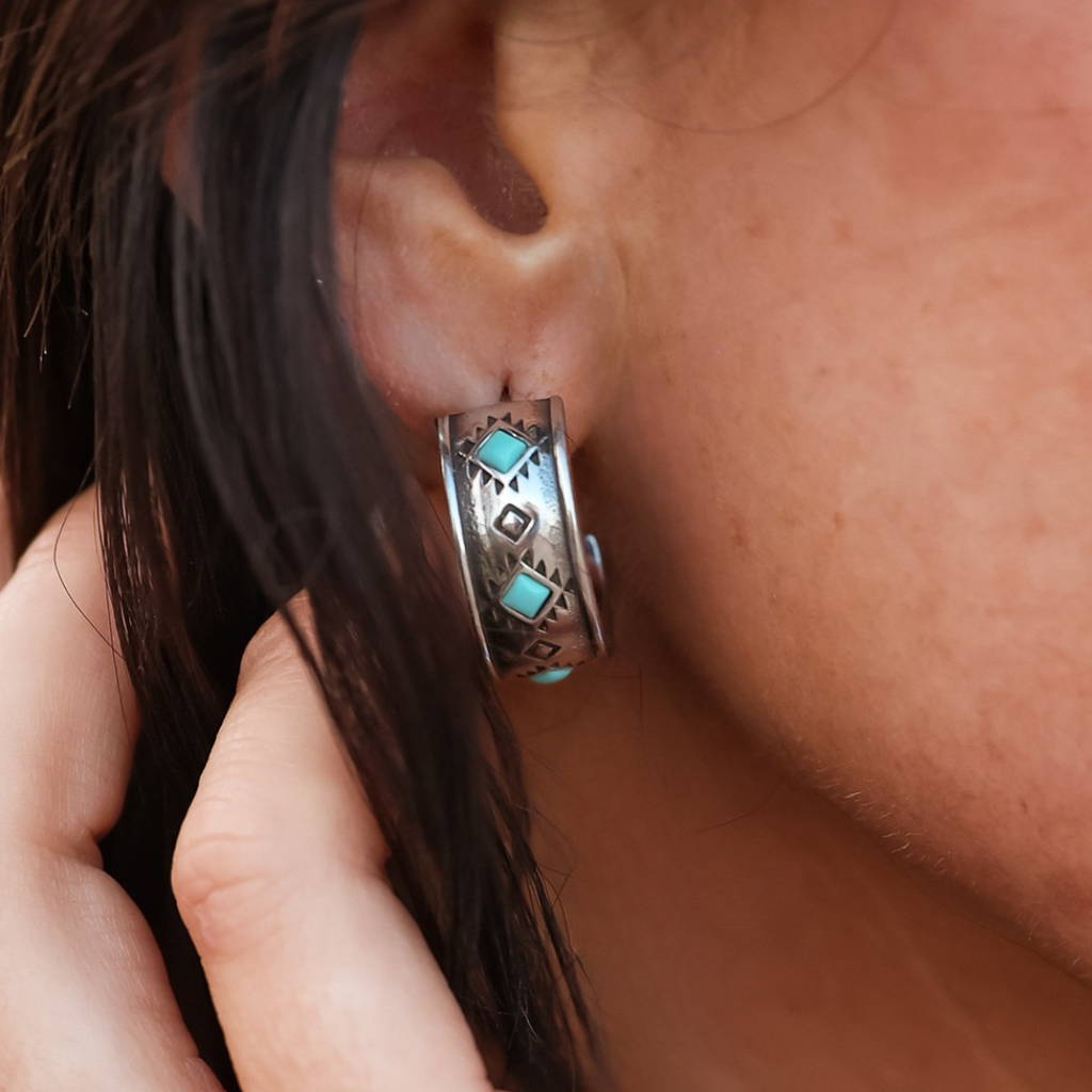 A close-up of a person’s ear wearing The Atzi Hoops—silver hoop earrings featuring turquoise geometric inlays and intricate etched designs that capture the essence of western jewelry. A hand is touching the hair near the ear.