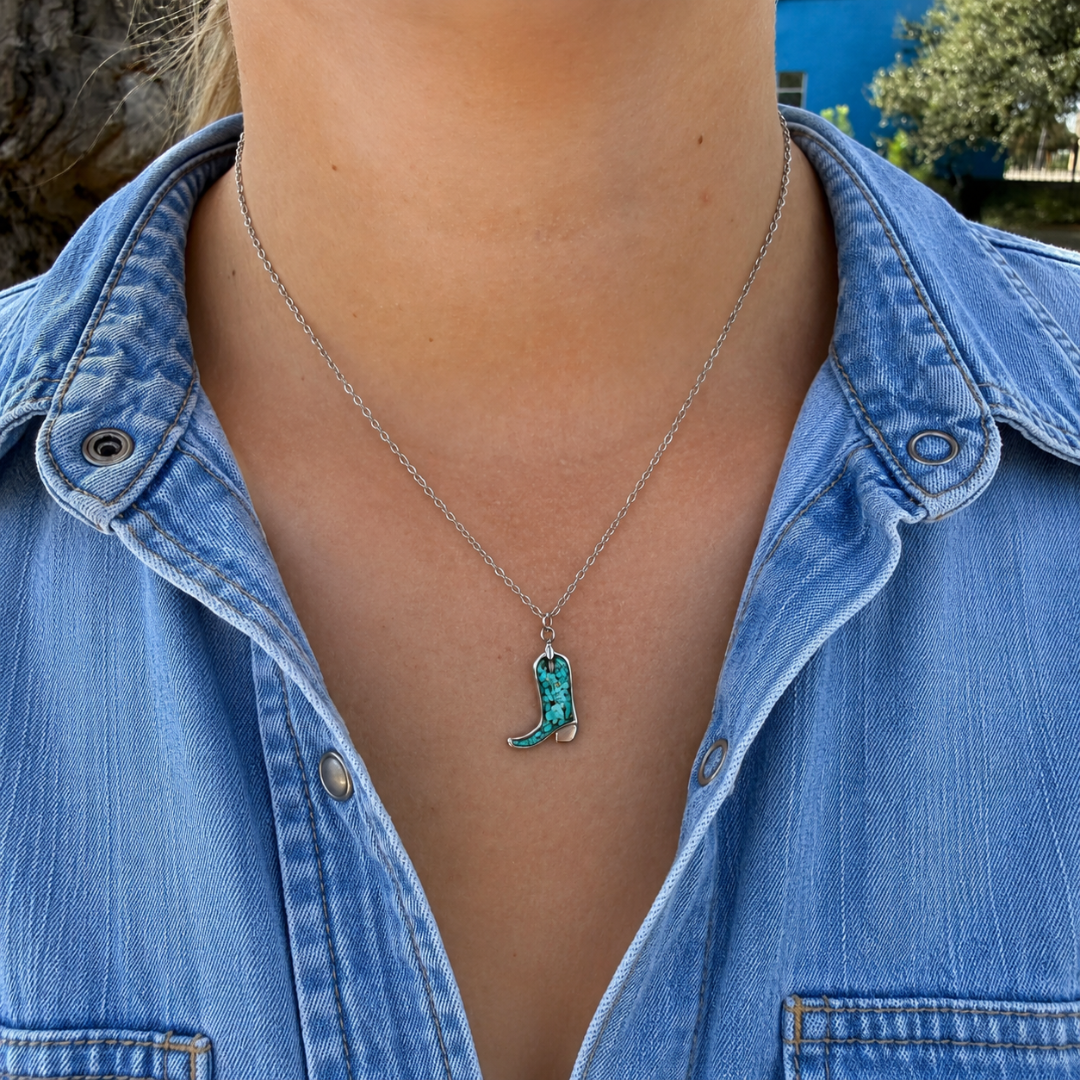 A person wearing The Boot Necklace—a silver chain necklace with a turquoise chili pepper pendant—styled with other turquoise jewelry and a blue denim shirt, standing outdoors.