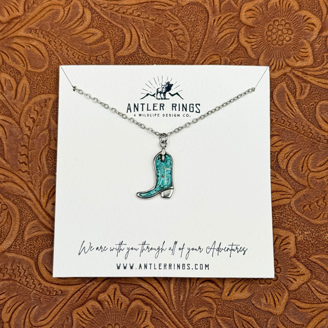 Introducing The Boot Necklace: a turquoise cowgirl boot pendant on a silver chain, beautifully displayed on an Antler Rings branded card against a brown tooled leather background—an eye-catching piece of turquoise jewelry and the ideal necklace for any western-inspired style.