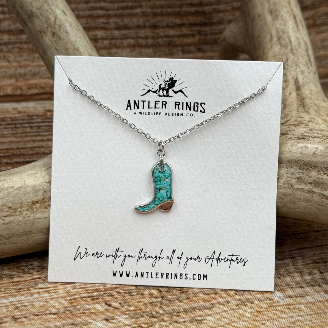 The Boot Necklace is showcased on an Antler Rings card, set against a rustic background of antlers and wood. The card features the Antler Rings website and a handwritten-style message about adventures, making The Boot Necklace the perfect addition to your turquoise jewelry collection.