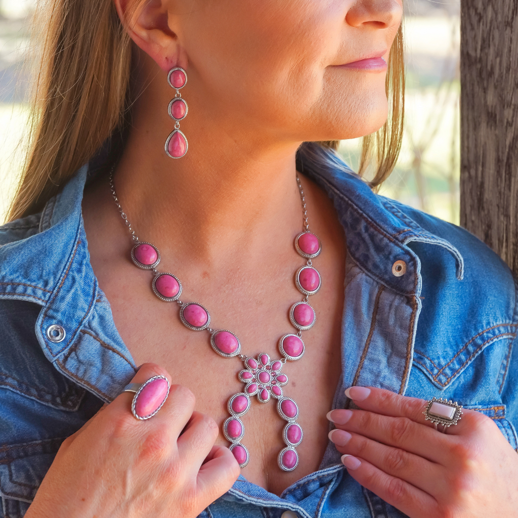 A woman in a denim shirt wears a matching set of The Charlie pink rhodonite jewelry—including a necklace, earrings, a ring, and a rectangular ring on her other hand. She stands by a wooden post, looking to the side and gently holding her shirt collar.