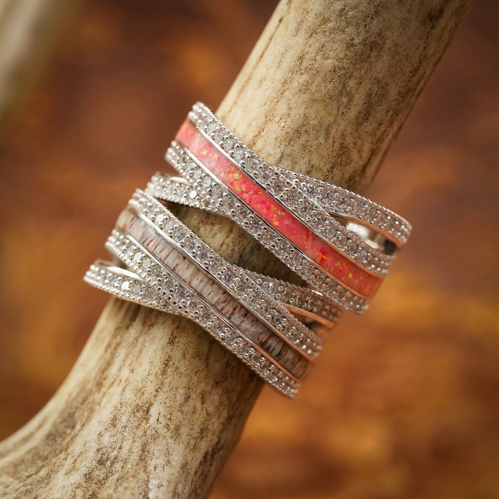The Chelly, a multi-band sterling silver ring featuring red, pink, and white inlays with sparkling stones, is wrapped around an antler against a blurred brown background.