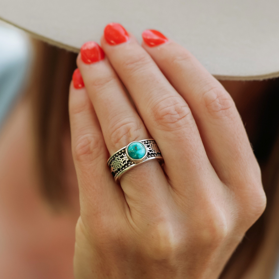 A hand adorned with red nail polish wears The Christoval—a sterling silver ring featuring intricate designs and a round turquoise stone—perfect for stacking or pairing with antler rings.