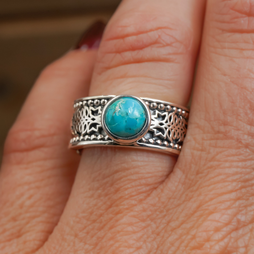 A close-up of a hand wearing The Christoval, a sterling silver ring featuring intricate patterns and a round turquoise stone at its center, beautifully paired with stacking rings for an elegant, layered look.