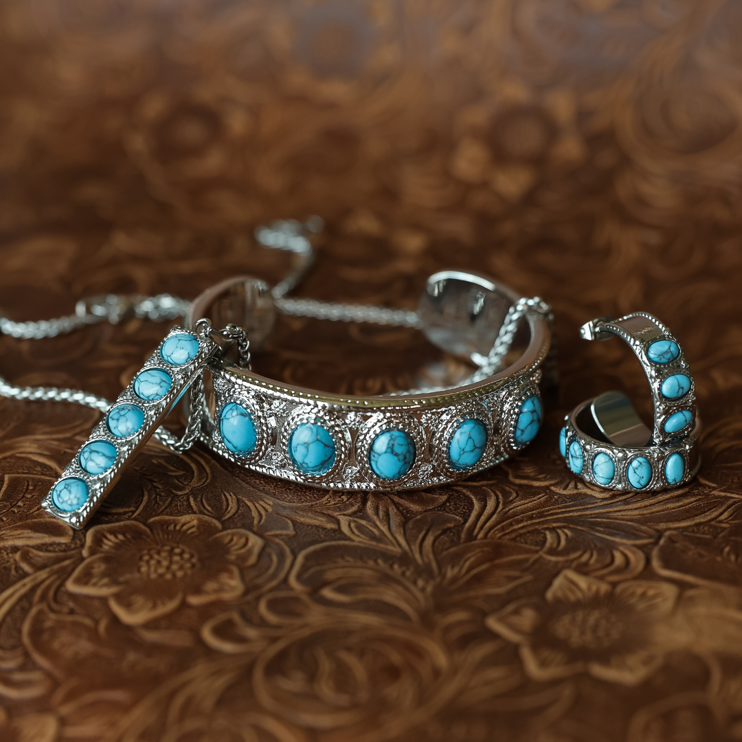 A silver-toned jewelry set featuring a cuff bracelet, a matching drop pendant, and a pair of hoop earrings, all embellished with oval turquoise stones, displayed on a textured brown leather background.