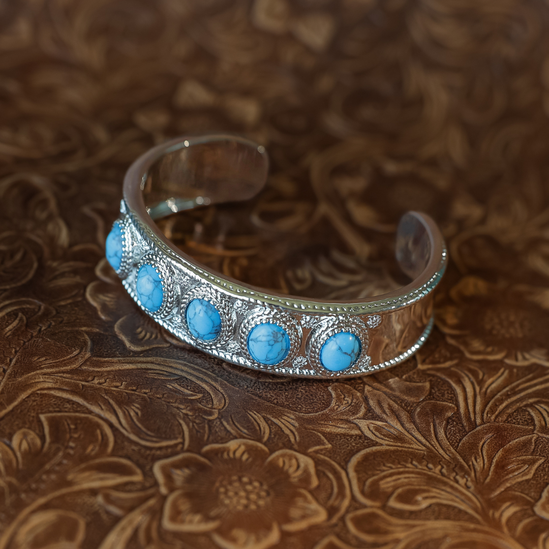 Introducing The Clara Cuff—a striking Western bracelet crafted in silver, featuring five round turquoise stones set against a brown, embossed floral leather background. Perfect for those who appreciate statement jewelry.