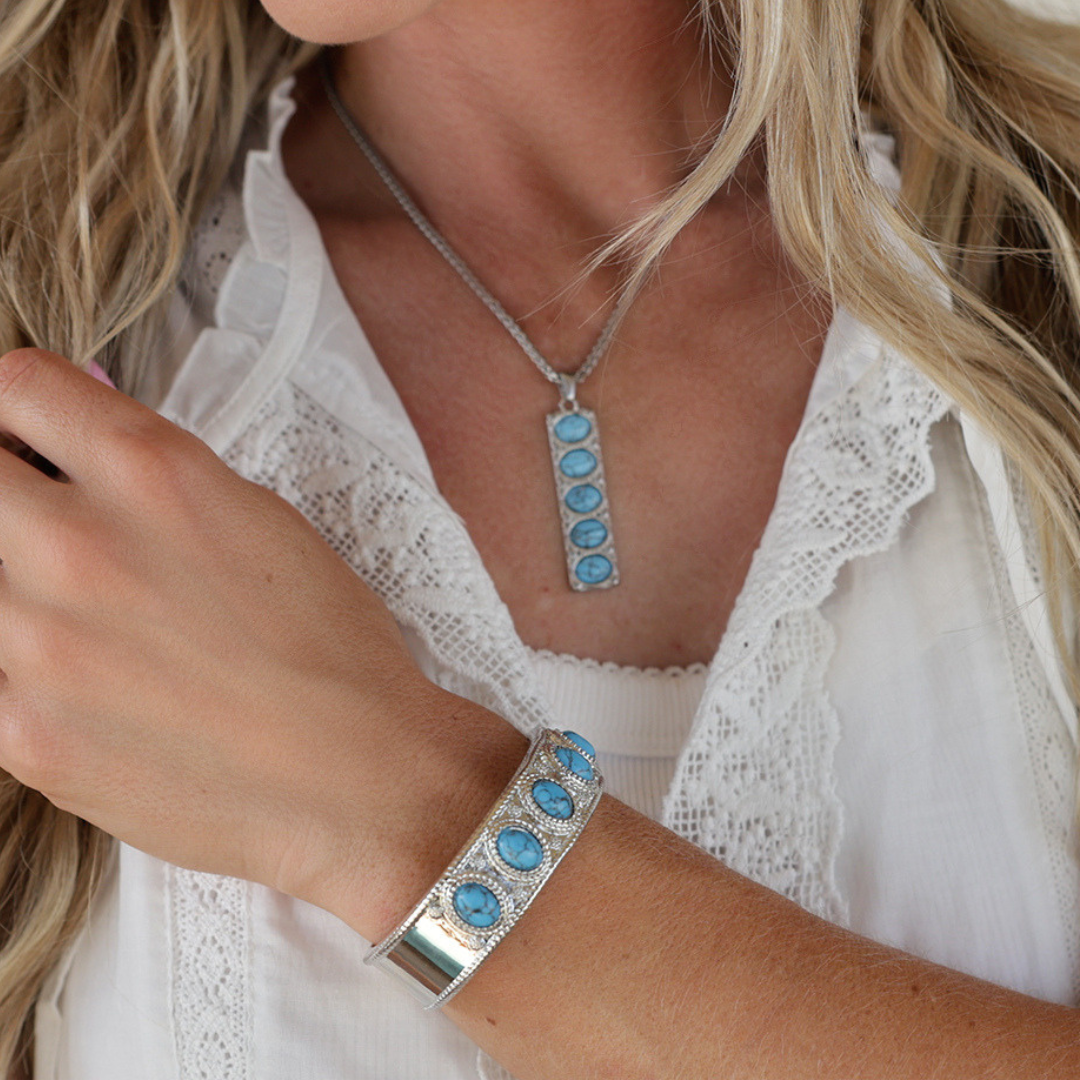 A woman wearing a white lace blouse showcases The Clara Cuff, a striking turquoise bracelet adorned with bold turquoise stones, along with a matching necklace. With only her lower face and upper body visible, the statement jewelry pieces take center stage as she gently touches her arm.