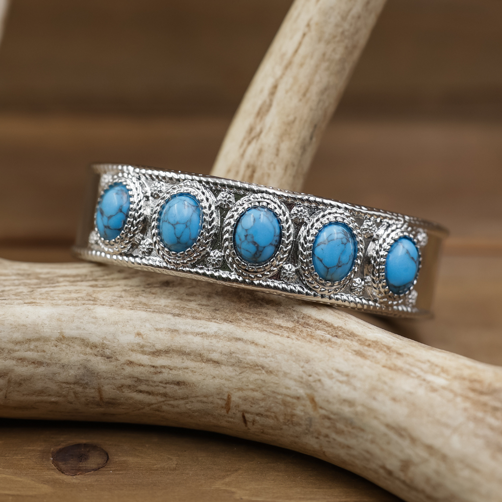 The Clara Cuff features five striking turquoise oval stones set in intricate silver metalwork, elegantly displayed on light-colored antlers against a blurred brown backdrop. This Western-inspired cuff makes a bold statement, seamlessly blending tradition with modern style.