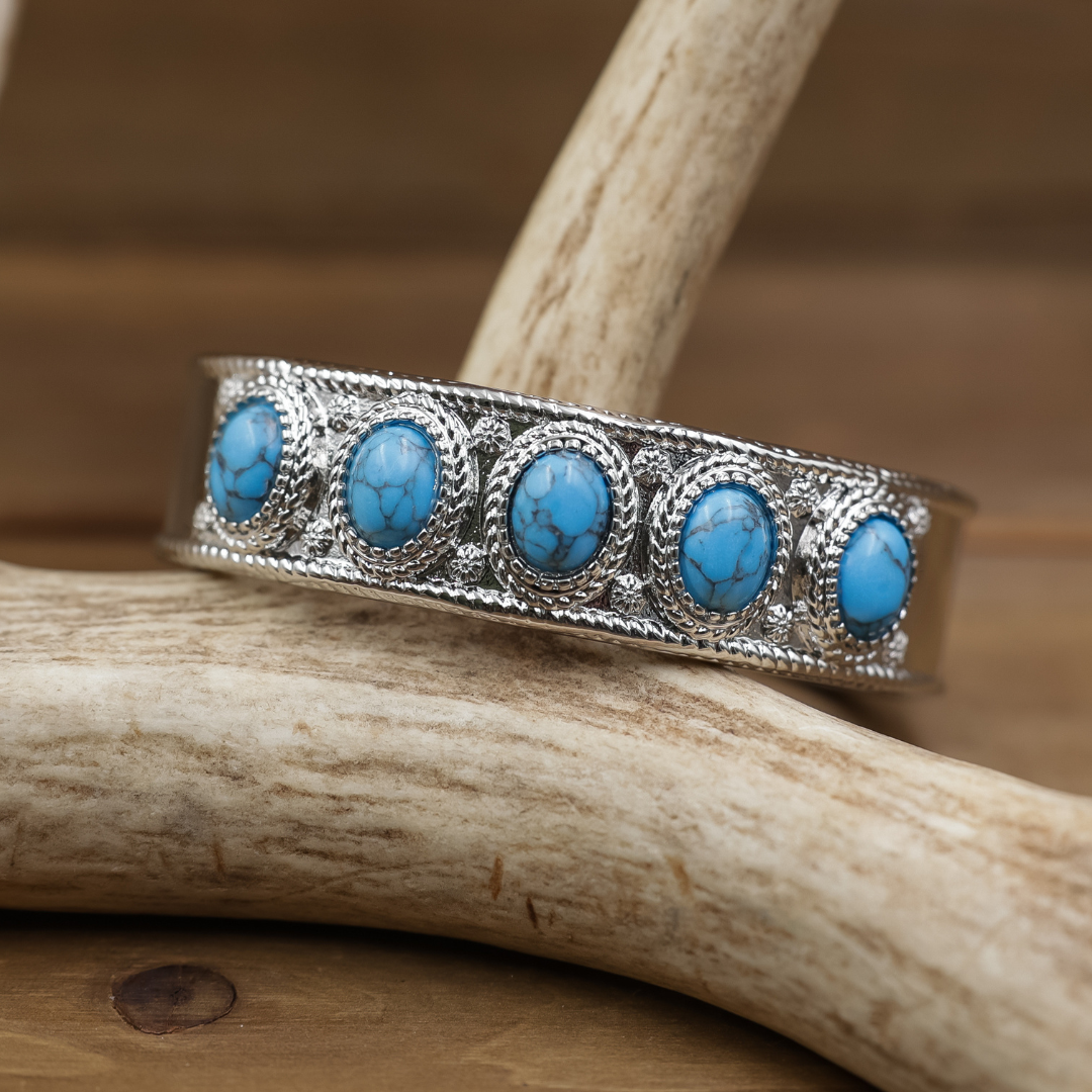 The Clara Cuff features five striking turquoise oval stones set in intricate silver metalwork, elegantly displayed on light-colored antlers against a blurred brown backdrop. This Western-inspired cuff makes a bold statement, seamlessly blending tradition with modern style.