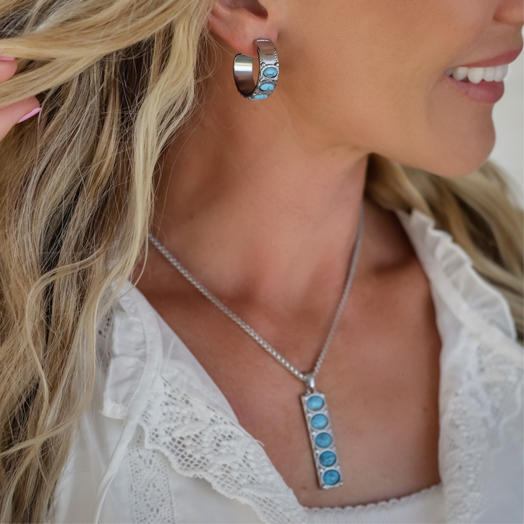 A close-up lifestyle shot of a woman wearing a matching silver-toned jewelry set, featuring a vertical bar pendant necklace and a hoop earring, both embellished with oval blue stones, paired with a white lace top.