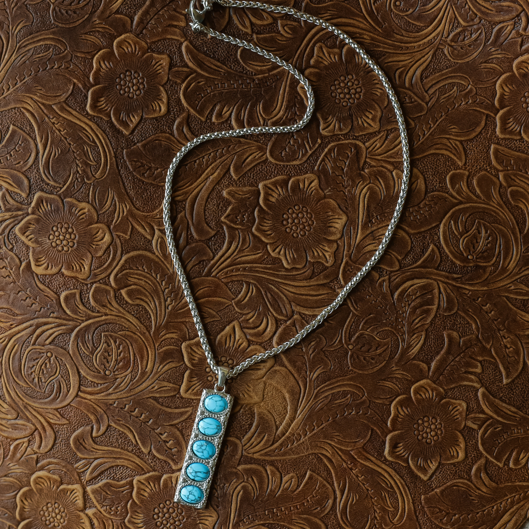 The Clara is a silver Western necklace with a rectangular pendant adorned with five turquoise stones, presented on embossed brown leather with a floral pattern.