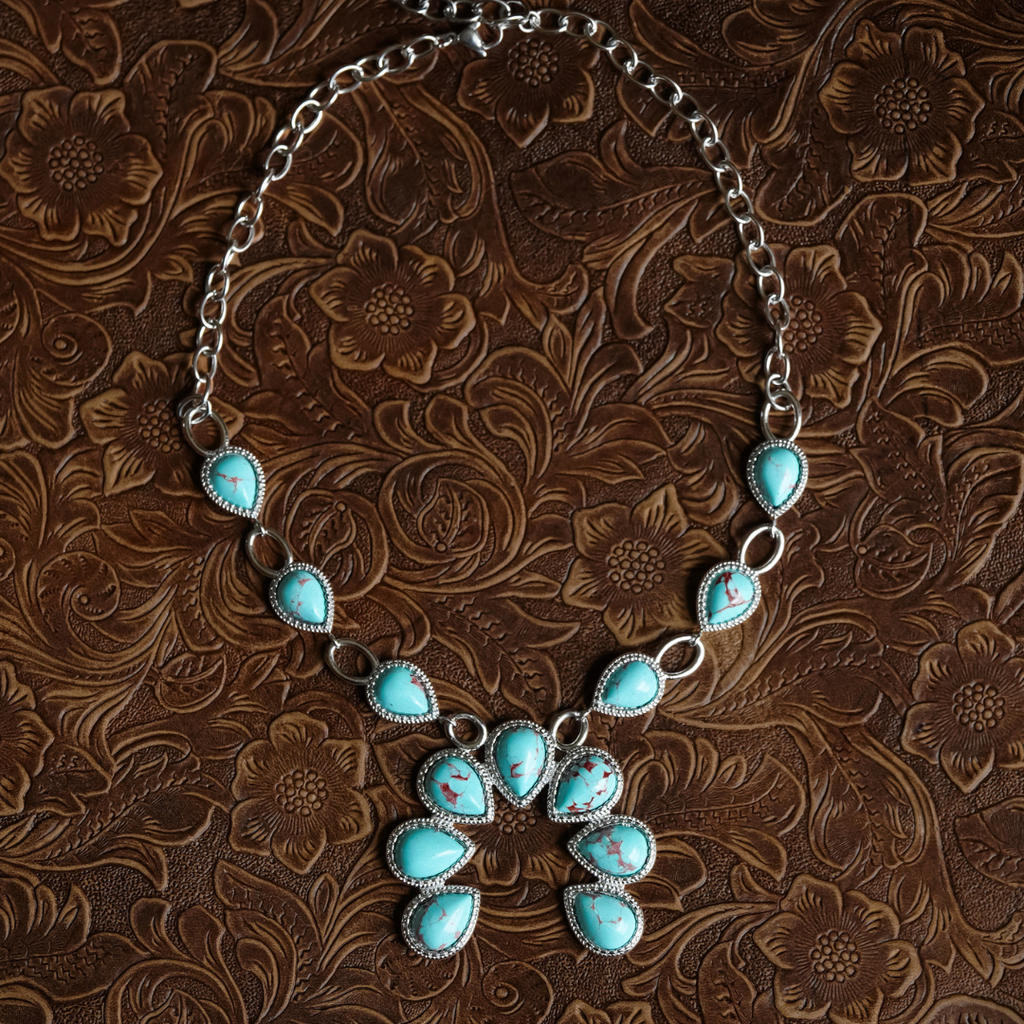 The Cornelia necklace, featuring silver Western design with turquoise stones in teardrop and oval shapes forming a floral-inspired pendant, is displayed on an embossed brown leather surface with a floral pattern.