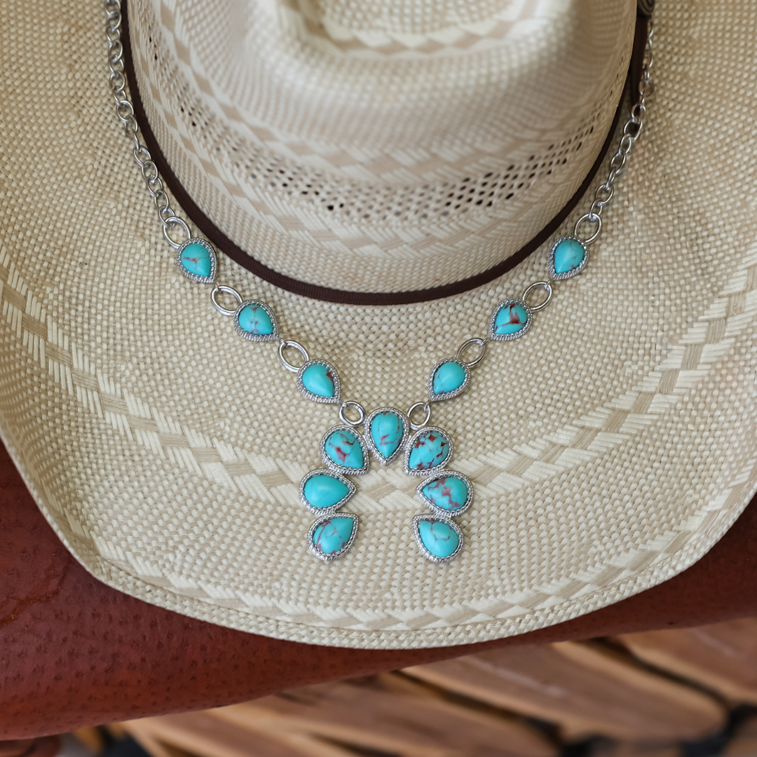 The Cornelia, a silver Western necklace adorned with turquoise teardrop and oval-shaped stones arranged in a floral pattern, is displayed on the front of a woven straw cowboy hat—perfectly blending engraved jewelry charm with Southwestern style.