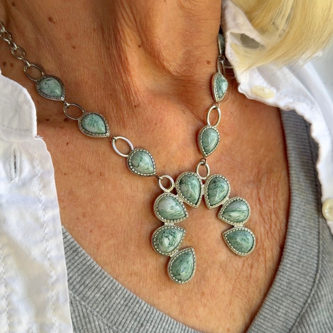 A person wearing a gray top and white shirt displays The Cornelia, a Western necklace featuring multiple turquoise teardrop-shaped stones arranged in a floral pattern, creating an eye-catching piece of engraved statement jewelry.