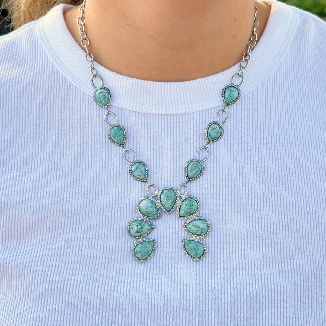A person wearing The Cornelia, a silver Western necklace featuring multiple green turquoise stone pendants, layered over a white ribbed shirt.