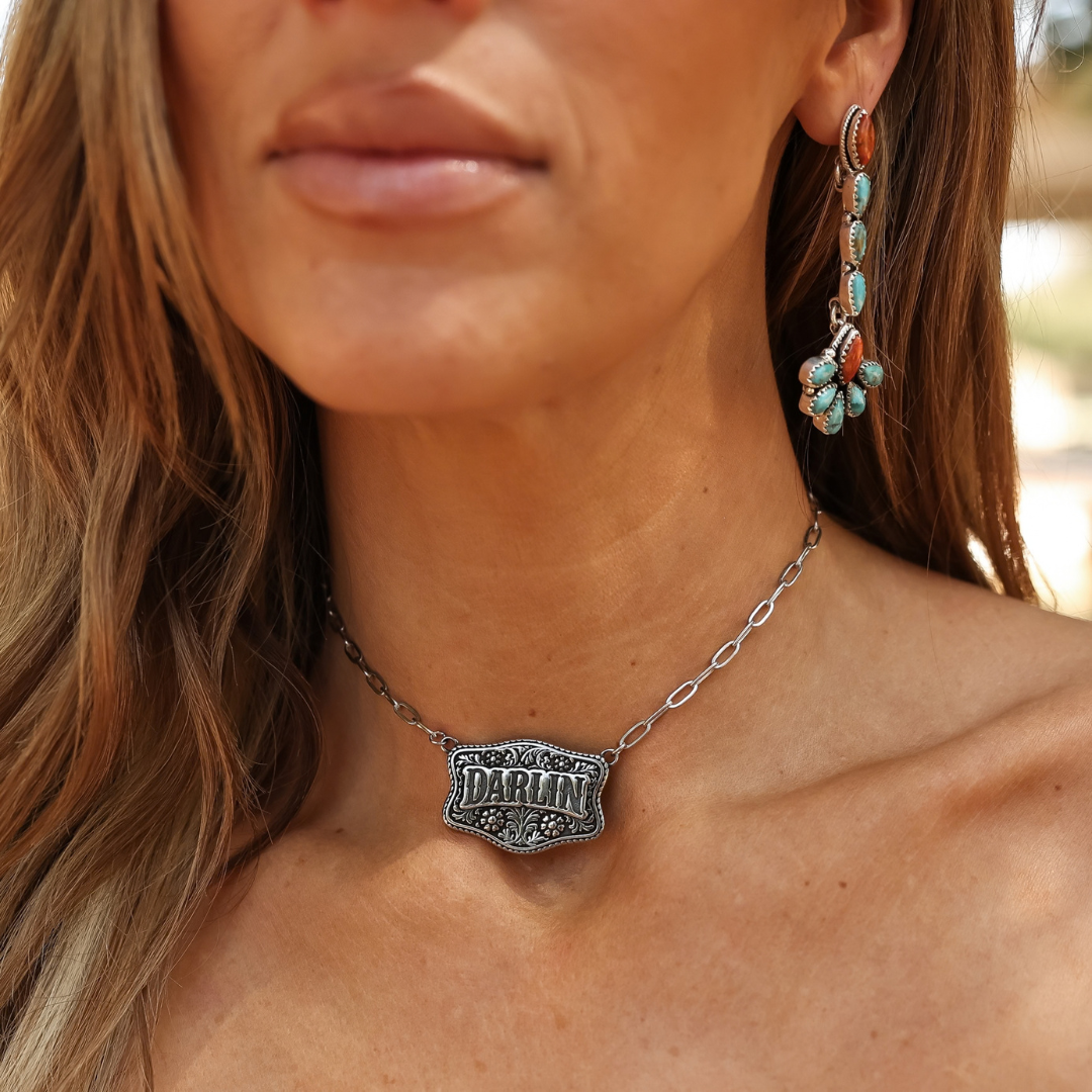 The Darlin’ silver statement necklace features a rectangular, floral engraved pendant, worn with matching ornate turquoise and coral drop earrings.