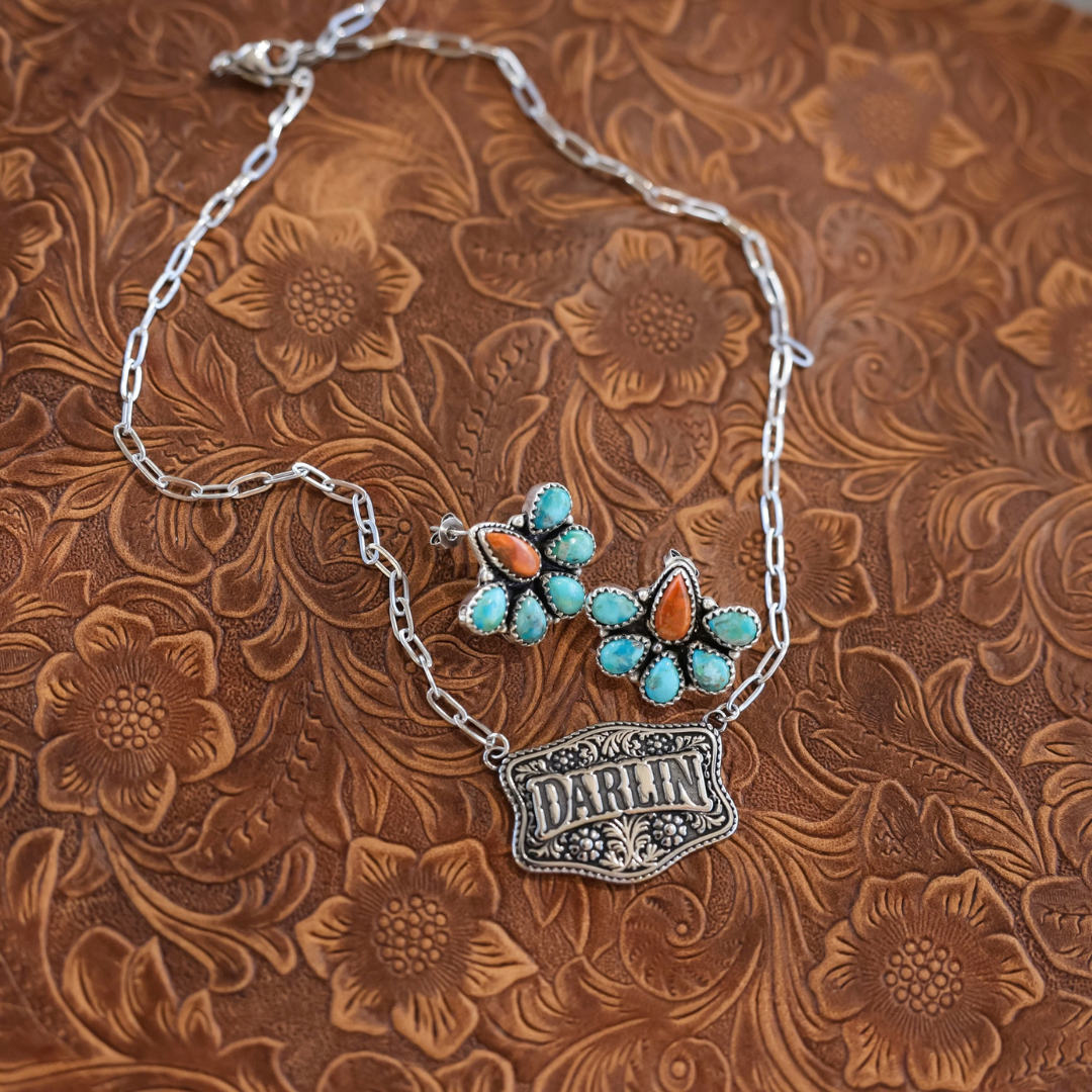 The Darlin’ silver floral engraved necklace featuring a rectangular pendant with the word DARLIN and a pair of matching flower-shaped stud earrings, each set with turquoise and orange stones.