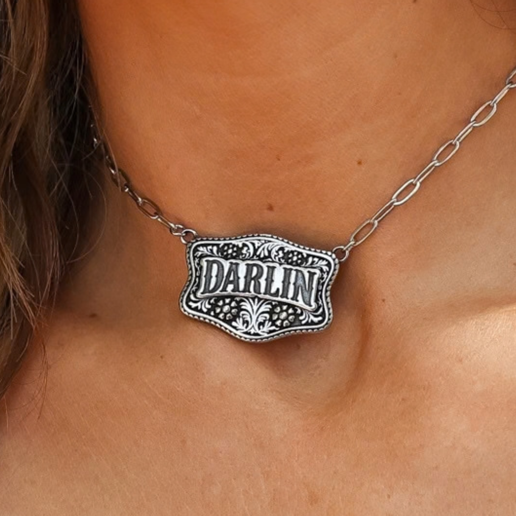 The Darlin’ silver-toned western necklace featuring a floral engraved rectangular pendant with the word DARLIN in the center on a metal chain.