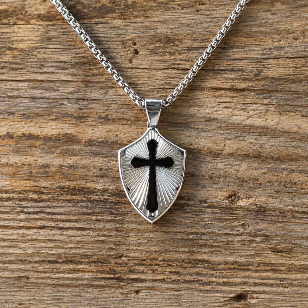 The David is a silver-tone metal shield necklace featuring a black cross, hanging from a chain against a wood background.