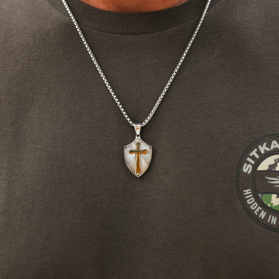 The David is a silver chain necklace with a shield-shaped cross pendant featuring a gold-toned cross at its center.