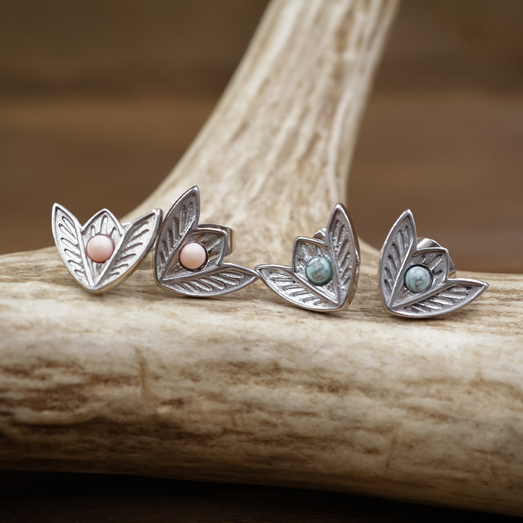 Two pairs of The Dessie Stud Earrings, crafted in the style of Western stud earrings with a silver leaf-shaped design, each feature a round gemstone—one pink, one blue-green—displayed atop a piece of antler against a softly blurred brown background. The blue-green stones evoke the timeless appeal of turquoise earrings, while the robust build suggests premium stainless steel quality.