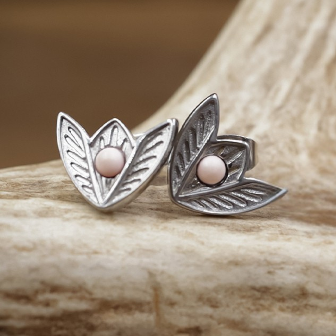 The Dessie Stud Earrings are a pair of silver Western-style studs shaped like leaves with delicate vein details, each adorned with a small round pink bead at the center, displayed on a textured beige surface.