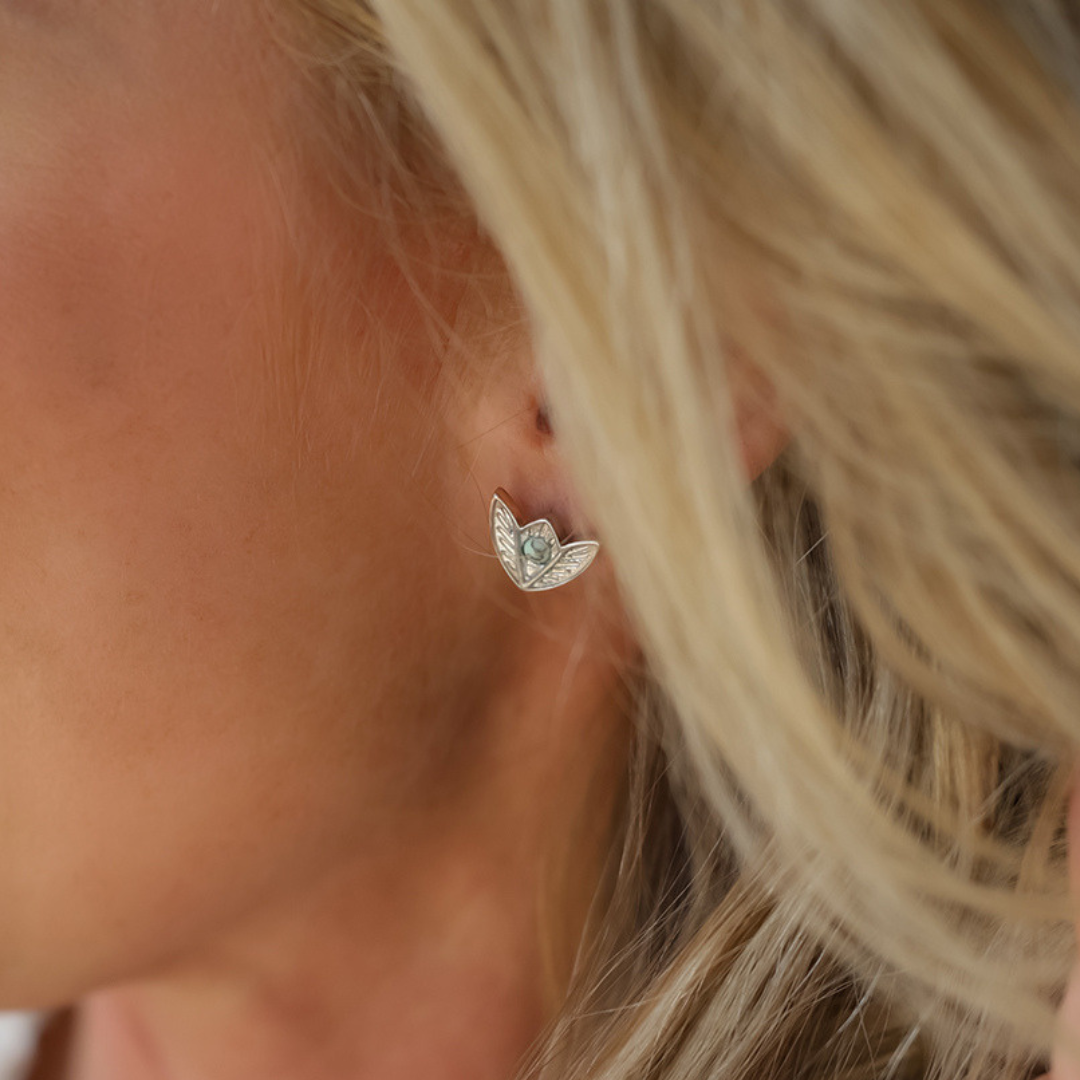 A close-up shows a person’s ear adorned with The Dessie Stud Earrings—Western-inspired studs shaped like angel wings and featuring a small, light blue faux turquoise stone at the center. The person’s blonde hair serves as a backdrop, accentuating the elegant stainless steel design of these eye-catching earrings.