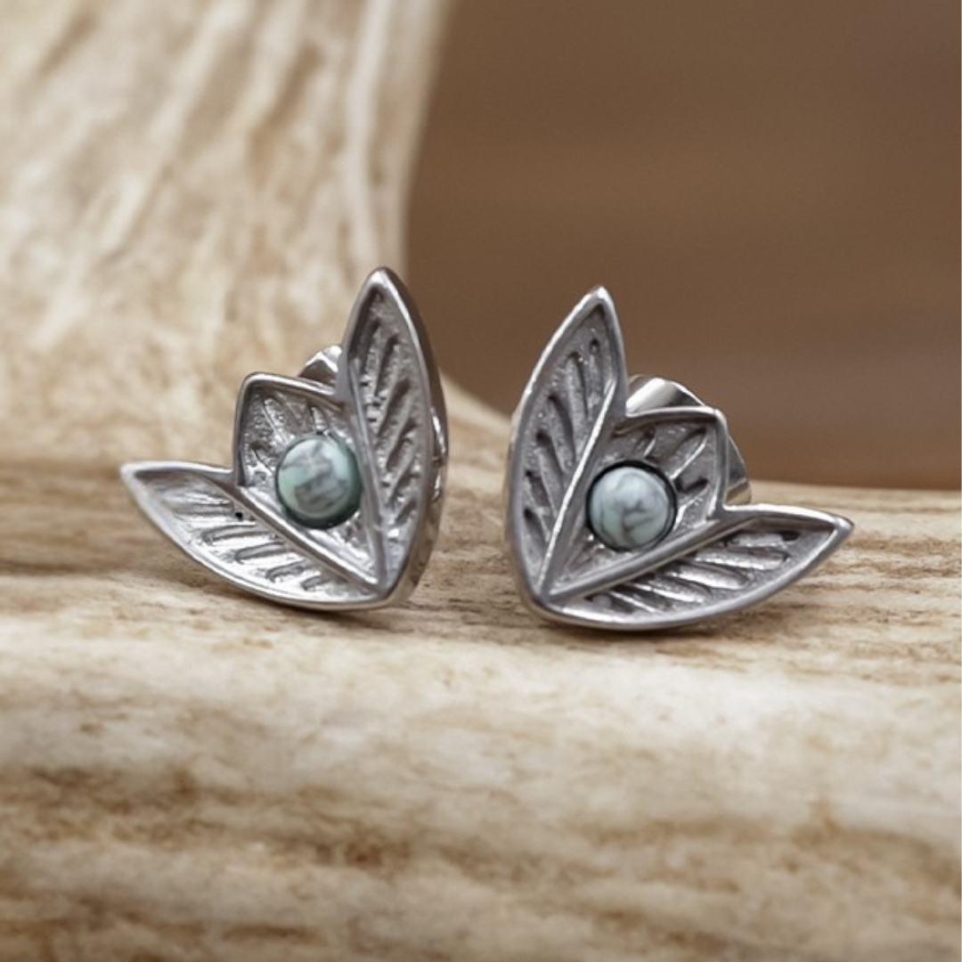 The Dessie Stud Earrings feature a pair of silver, leaf-shaped Western studs adorned with green marbled stones at the center, displayed on a light wooden surface.