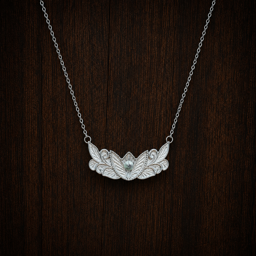 The Dessie—a silver floral necklace with an ornate leaf and lotus flower design, featuring a small turquoise pendant in the center—is displayed on a dark wood background.