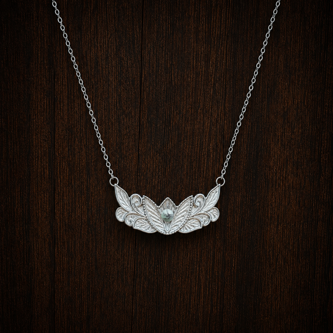 The Dessie—a silver floral necklace with an ornate leaf and lotus flower design, featuring a small turquoise pendant in the center—is displayed on a dark wood background.