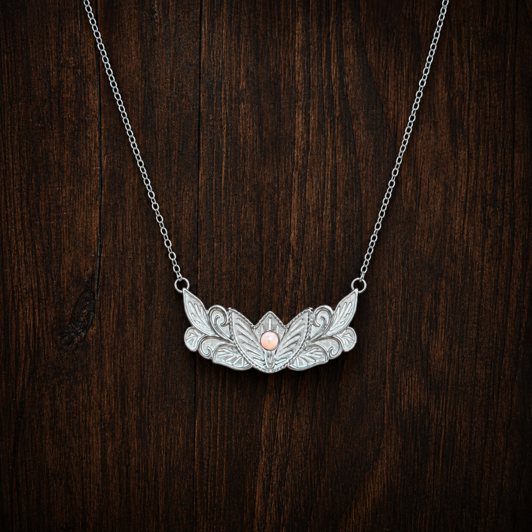 The Dessie, a silver Western necklace featuring an ornate leaf pattern with a single pink pearl at its center, is showcased against a dark wooden background and makes a stunning addition to your floral jewelry collection.