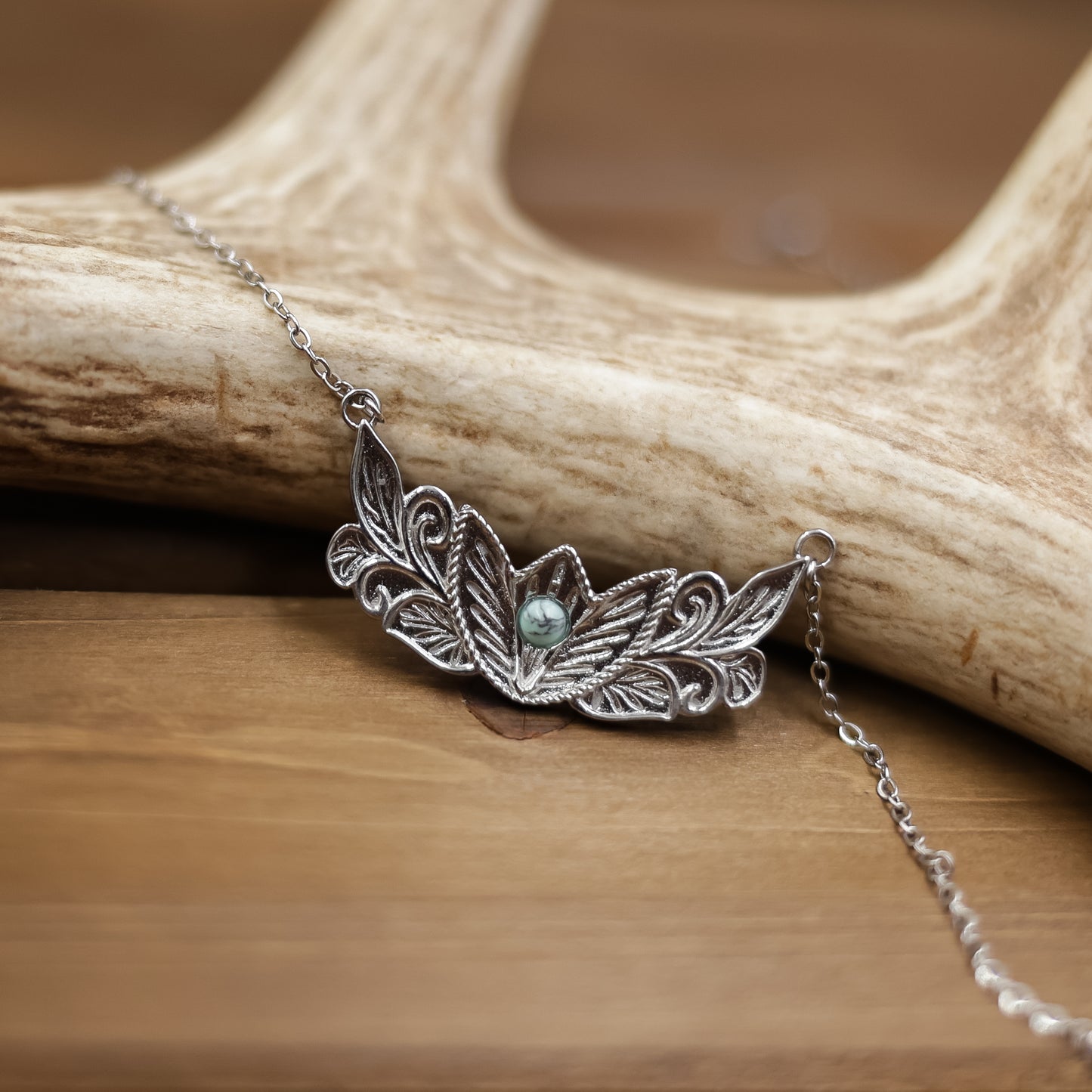 The Dessie, a silver Western necklace featuring an ornate leaf design and a small turquoise stone at its center, is displayed draped over an antler on a wooden surface.
