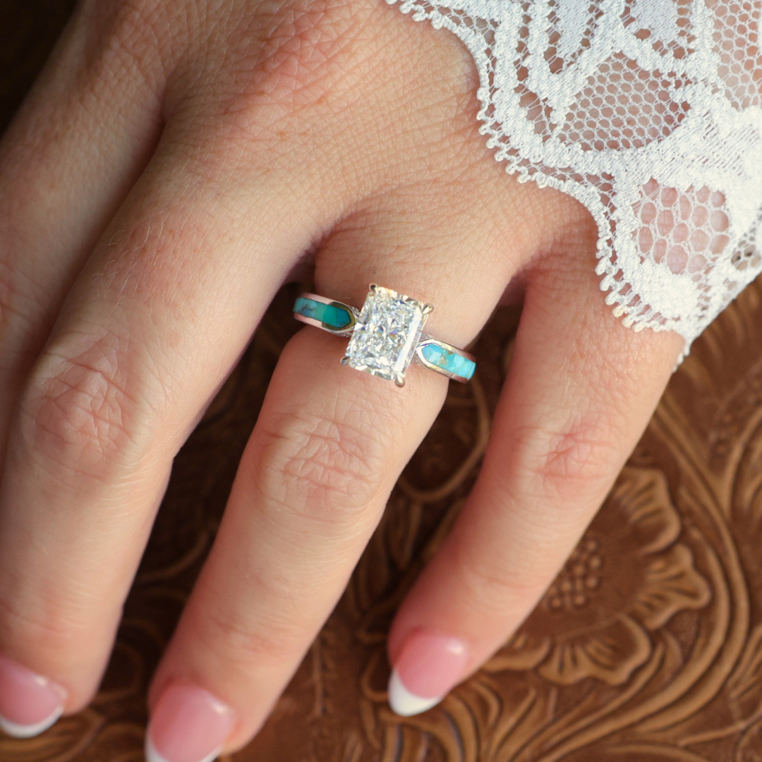A hand with a French manicure wears the Diamond Rochelle, a silver, Western-inspired engagement ring featuring a rectangular clear gemstone and striking turquoise inlay, partially covered by delicate white lace fabric.