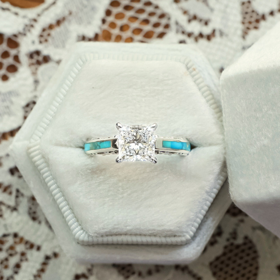 The Diamond Emory, a Western-inspired engagement ring with a silver band, showcases a square-cut lab-grown diamond and vibrant turquoise inlays. It is presented in a white, hexagonal velvet ring box atop lace fabric.