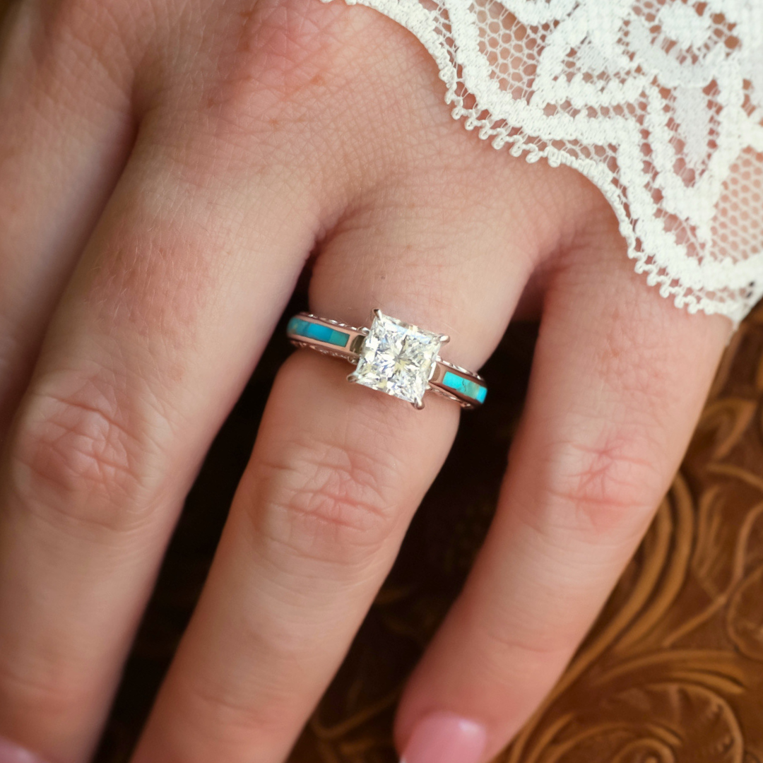 A hand adorned with the exquisite Diamond Emory lab-grown diamond engagement ring, featuring a turquoise inlay band, rests gracefully on a brown surface. A delicate lace accent near the fingers enhances the elegance and Western-inspired charm of this remarkable piece.