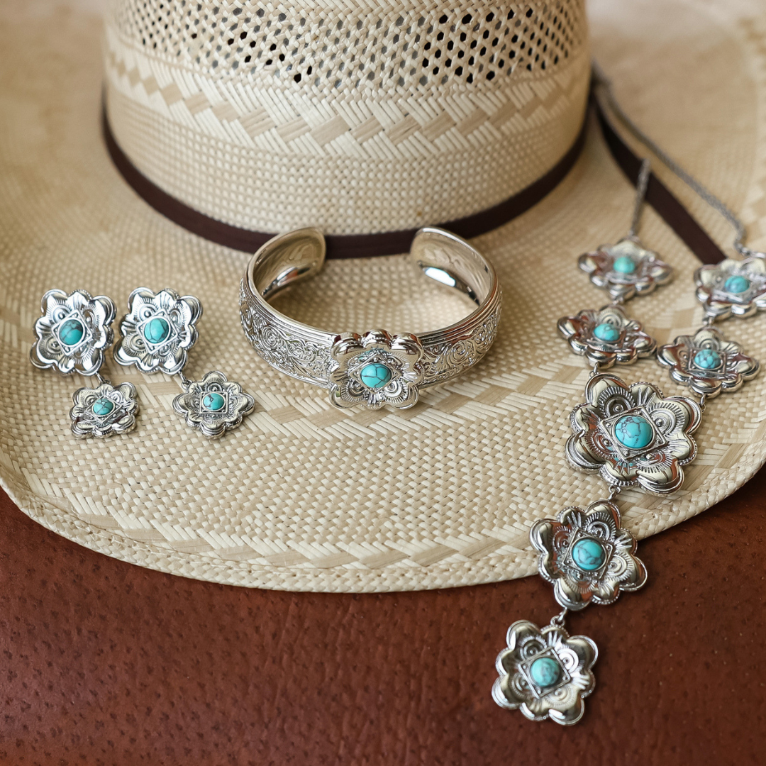 A straw cowboy hat displays turquoise and silver jewelry, including The Eloise Drop Earrings, a bracelet, and pendants—all arranged on a brown leather surface.