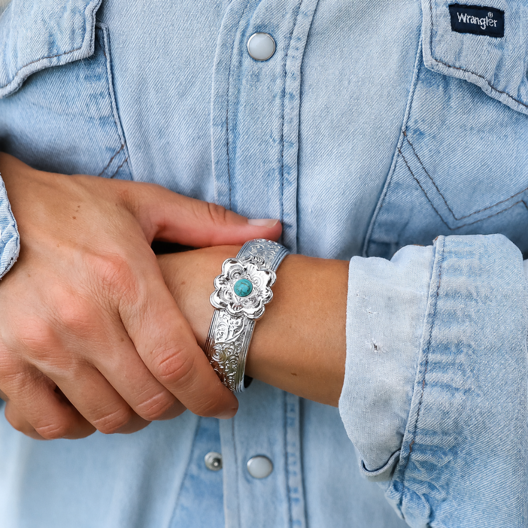 Wearing a light blue denim shirt, a person holds their wrist to showcase The Eloise Cuff—a silver Western cuff adorned with an engraved floral design and featuring a striking turquoise stone at its center, making it a true statement piece.
