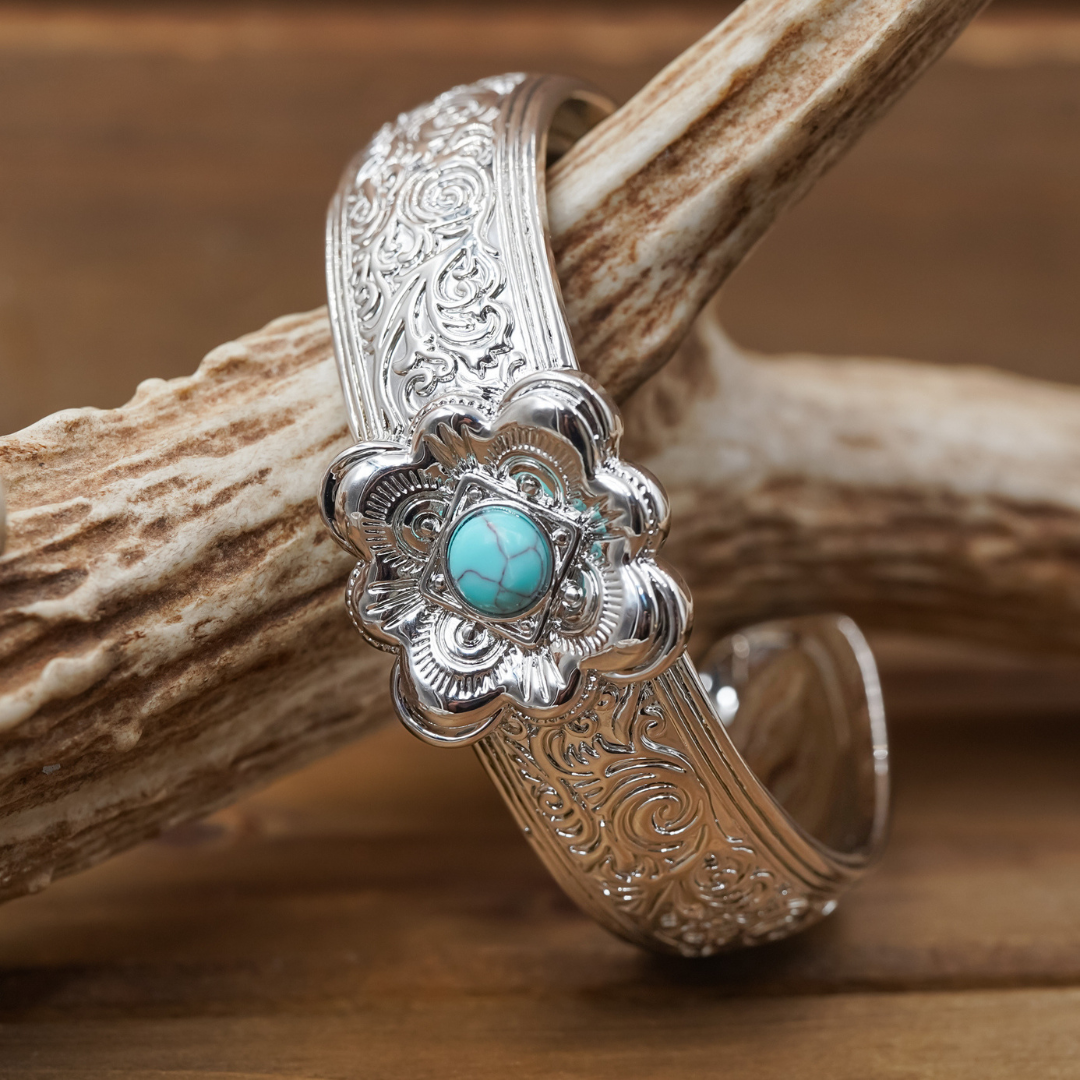 Introducing The Eloise Cuff: an ornate silver bracelet featuring intricate floral engravings and a vibrant blue turquoise stone centerpiece. Displayed on a textured wooden branch, this striking turquoise cuff is perfect for lovers of statement jewelry.