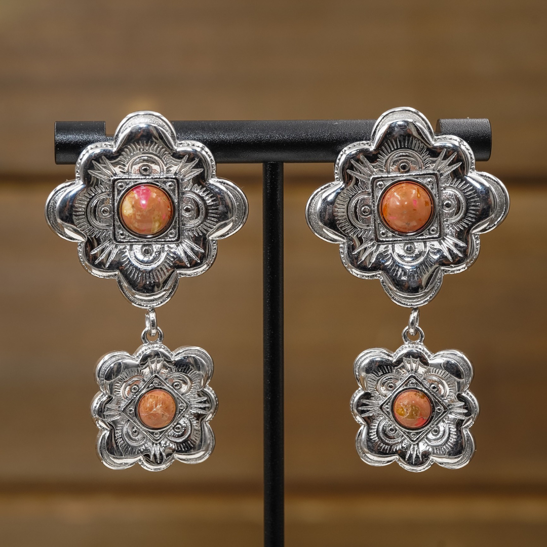 The Eloise Drop Earrings are stunning silver floral statement pieces featuring intricate designs, each adorned with a central orange gemstone and a smaller matching dangle. Displayed on a black stand against a blurred brown background, these Western-inspired earrings are perfect for elevating any look.