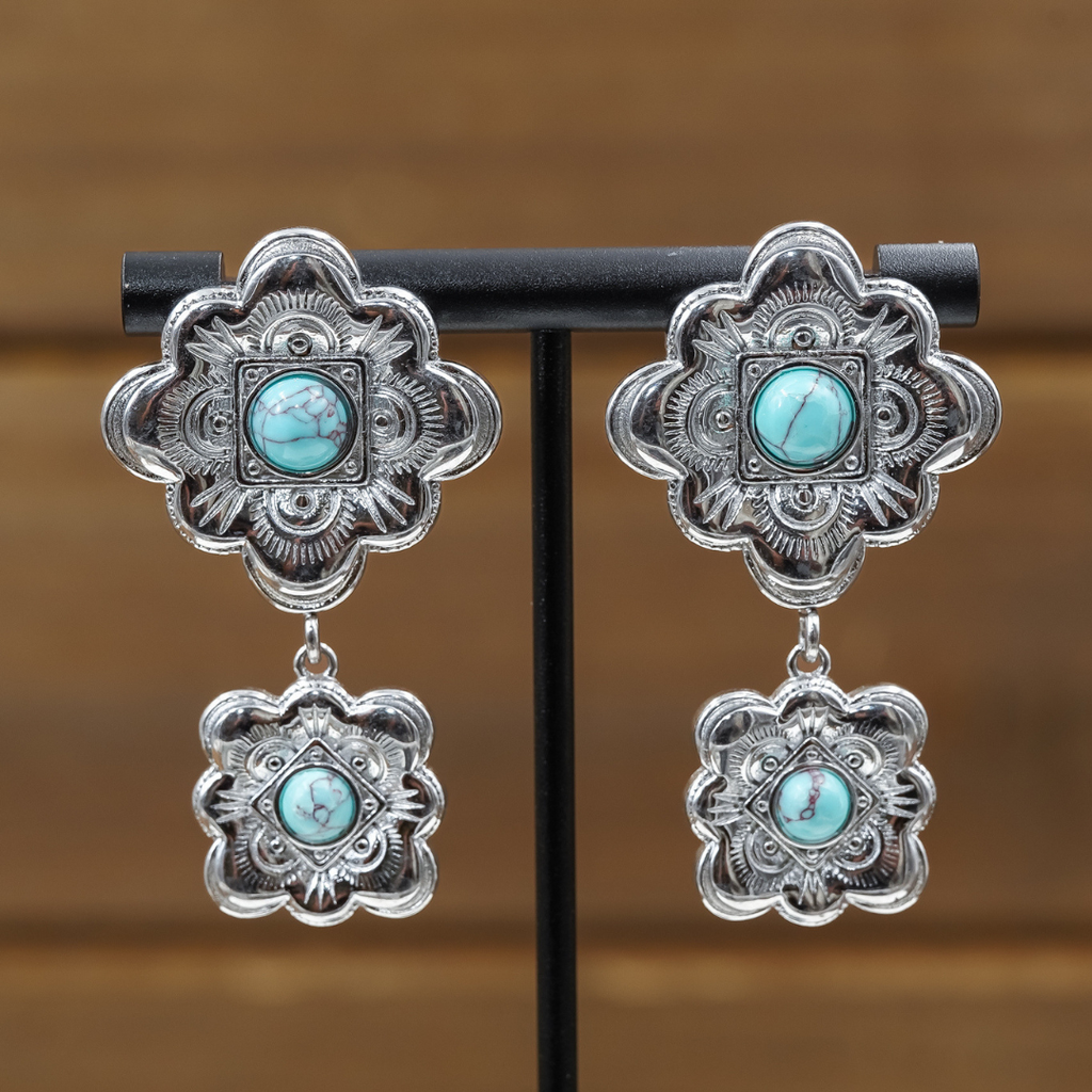Introducing The Eloise Drop Earrings: silver statement earrings adorned with intricate floral patterns, each showcasing a central turquoise stone and a coordinating, smaller drop piece below. These striking turquoise earrings are presented on a black stand set before a softly blurred brown backdrop.