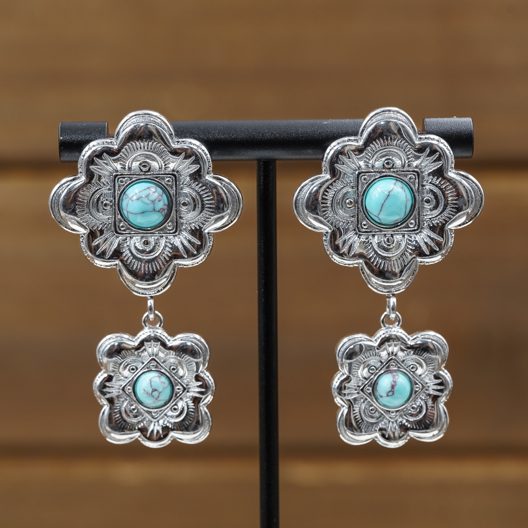 Introducing The Eloise Drop Earrings: silver statement earrings adorned with intricate floral patterns, each showcasing a central turquoise stone and a coordinating, smaller drop piece below. These striking turquoise earrings are presented on a black stand set before a softly blurred brown backdrop.