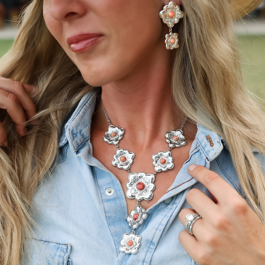 Wearing a denim shirt, a woman models The Eloise, a heritage-inspired jewelry set featuring coral-colored floral designs. The set includes earrings, a Western necklace, and a ring.