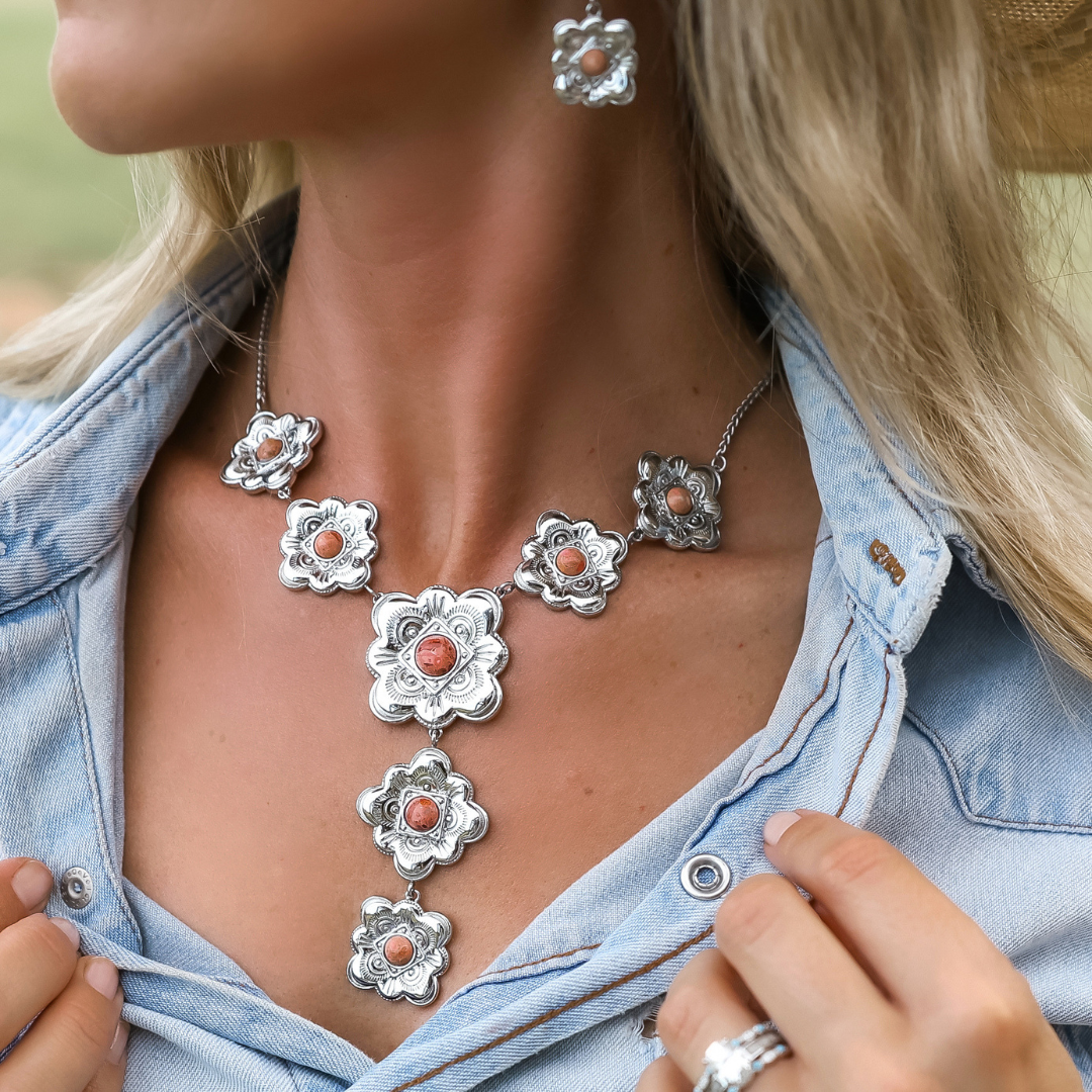 Woman wearing a denim shirt and The Eloise—a silver and coral floral necklace with matching earrings and ring—showcasing the heritage-inspired jewelry close to her neck and chest.