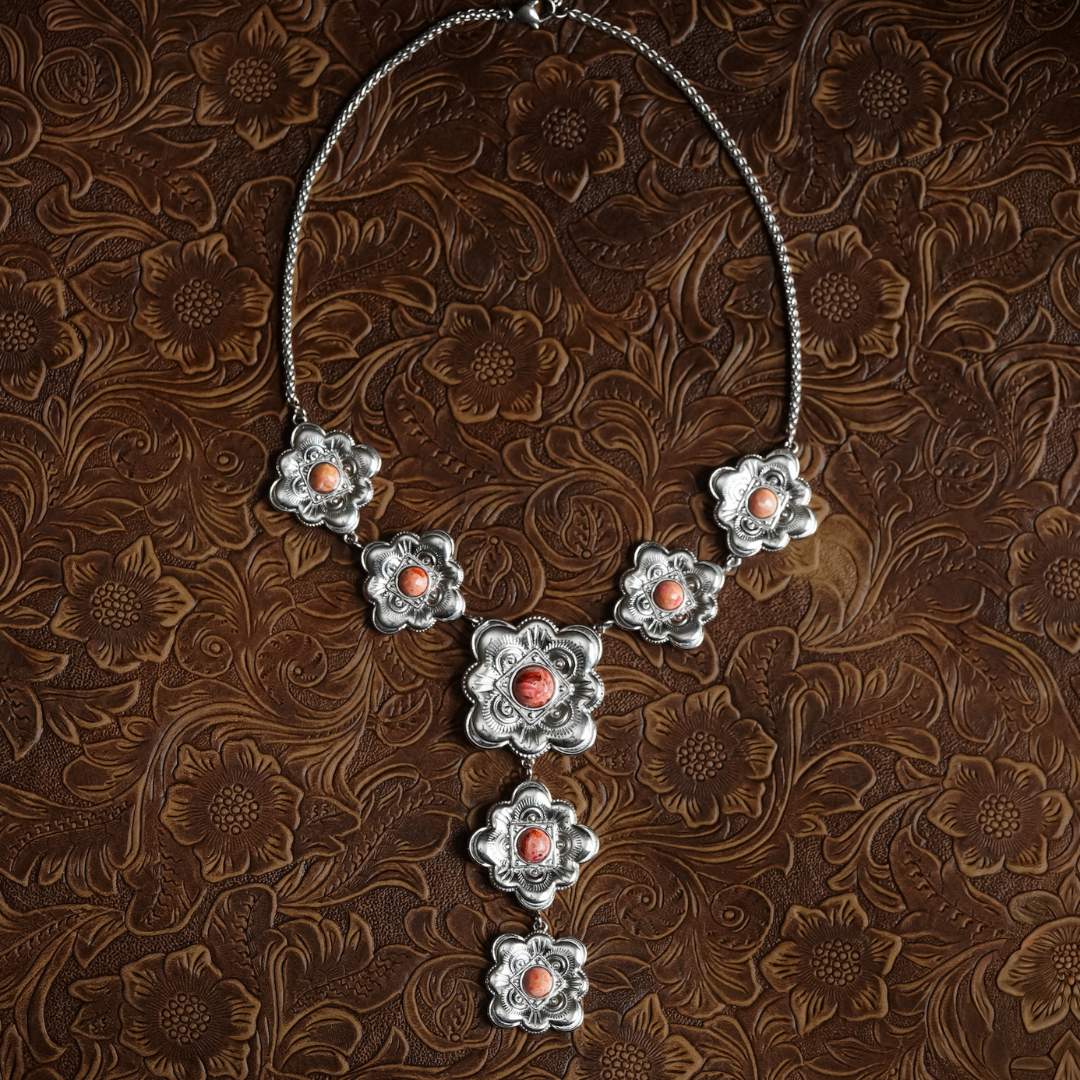 The Eloise is a silver floral necklace adorned with intricately crafted flower-shaped pendants, each set with a pink center stone. Displayed on an embossed brown floral background, The Eloise captures the charm of heritage-inspired jewelry, seamlessly blending timeless elegance with artisanal craftsmanship.