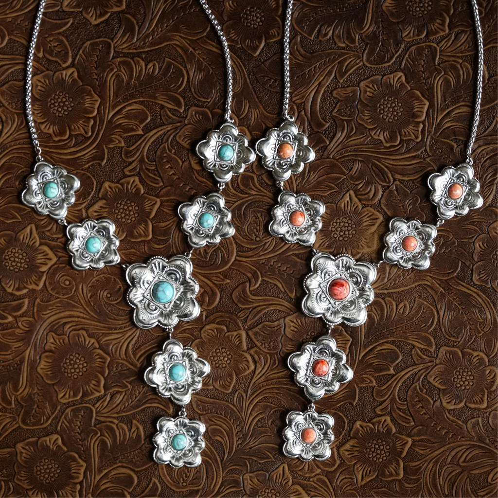 Two ornate silver necklaces—The Eloise—with flower-shaped pendants, each featuring blue-green turquoise or coral-colored stones, are displayed on a brown leather surface with a floral embossed pattern: perfect examples of heritage-inspired jewelry.