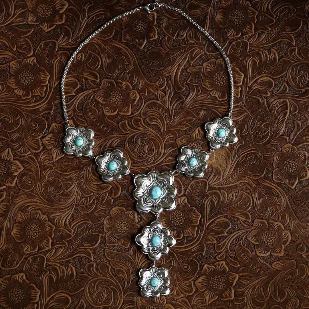 The Eloise is a silver Western necklace adorned with seven floral pendants, each set with a vibrant turquoise stone, showcased on brown embossed leather with an intricate floral pattern—a stunning piece of heritage-inspired jewelry.