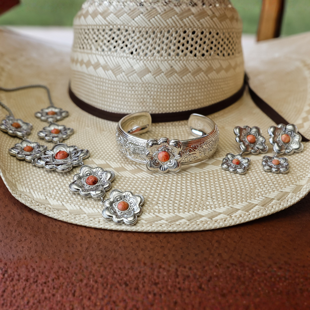 A straw cowboy hat displays The Eloise Drop Earrings—silver jewelry with floral designs and orange stones—alongside a bracelet, a ring, and a necklace, on a brown leather surface.